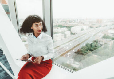 A beautiful African-American woman entrepreneur in a white blouse and red skirt is holding a digital tablet pc while sitting indoors on the bench on the top floor of a business office skyscraper